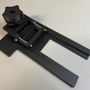 Può includere: Supporto per fotocamera in plastica nera stampato in 3D con una manopola per regolare l'angolo. Il supporto ha un'apertura rettangolare per un obiettivo della fotocamera e una fessura per una diapositiva.