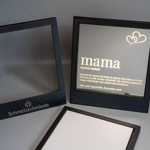 Könnte beinhalten: Zwei schwarze Bilderrahmen mit Glasfronten. Der Rahmen links hat einen leeren weißen Hintergrund. Der Rahmen rechts hat einen weißen Hintergrund mit goldener Schrift, die "mama" und "immeral women" lautet. Der Text setzt sich mit einer Definition des Wortes "mama" auf Deutsch fort. Zwei goldene Herzen befinden sich in der oberen rechten Ecke des Rahmens.