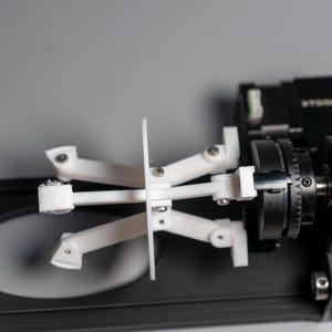 Könnte beinhalten: Ein weißer Roboterarm aus Kunststoff mit mehreren Gelenken und einem schwarzen Sockel. Der Arm befindet sich in einer angehobenen Position, wobei die Gelenke ausgefahren sind.