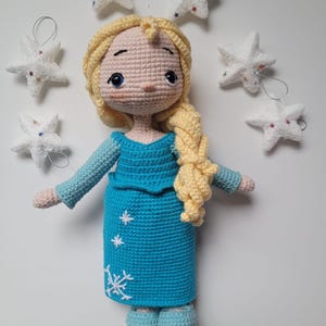 May include: A crocheted doll with long blonde hair wearing a blue dress with a snowflake design. The doll is surrounded by white crocheted stars.
