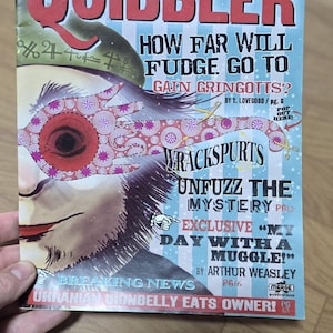 Peut inclure: Une copie du magazine "The Quibbler", avec une couverture colorée. Le titre est en rouge vif. La couverture présente des titres sur Fudge, Gringotts, Wrackspurts et une histoire d'Arthur Weasley. Le magazine est tenu par une main.