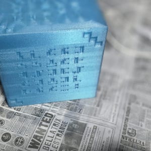 Könnte beinhalten: Ein hellblauer, strukturierter Würfel mit pixeligen Designs auf seinen Seiten. Der Würfel liegt auf einer Zeitung mit verschiedenen Schlagzeilen und Texten. Das Design des Würfels verleiht ihm eine digitale oder Retro-Ästhetik.