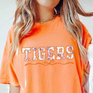 May include: Bright orange t-shirt with the word "TIGERS" in a floral design. The shirt has short sleeves and a crew neck. The text is in a bold, blocky font with a floral pattern inside the letters. The shirt is a comfortable, casual style.