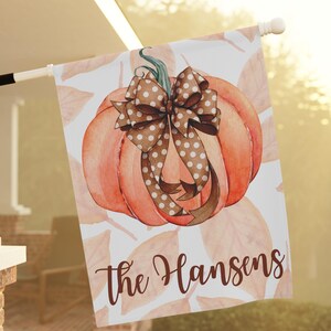 May include: A white house flag featuring a watercolor pumpkin with a brown polka dot bow. The pumpkin is orange with a green stem. The words "The Hansens" are written in brown script below the pumpkin. The background has a pattern of orange leaves.