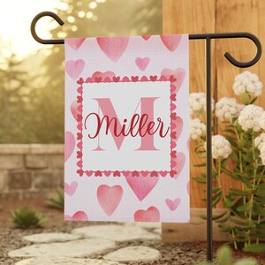 May include: A pink and white garden flag with a watercolour heart pattern. The flag has a white square in the centre with a large pink letter "M" and the word "Miller" in pink cursive writing.