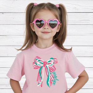May include: A pink t-shirt with a bow design, stripes, teal ribbons, and a pearl-like strand. The bow has the letter "P" and the name "Penelope". The girl is wearing heart-shaped sunglasses. The background is a white wooden wall.
