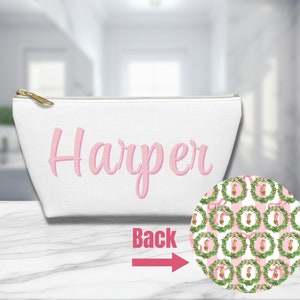 Puede incluir: Bolsa de maquillaje de lona blanca con una cremallera rosa y el nombre "Harper" bordado en rosa. La parte posterior de la bolsa presenta un patrón repetido de coronas verdes con cintas rosas.