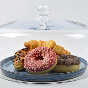 May include: A clear glass cake dome with a round handle covers a blue plate holding pastries. The plate contains a pink sprinkled donut, a chocolate frosted donut, and croissants. The dome sits on a clear glass base.