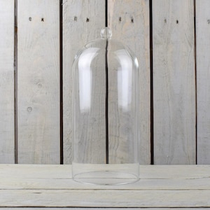 Może przedstawiać: Przezroczysta szklana klosz z zaokrąglonym wierzchołkiem i szeroką podstawą. Klosz stoi na białym drewnianym podłożu.