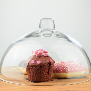 Może przedstawiać: Szklana kopuła z czekoladowym muffinkiem ozdobionym różowymi posypkami i różową lukrowaną pączkiem z białymi posypkami.