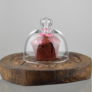Może przedstawiać: Czerwony welwetowy cupcake z różowymi posypkami w kształcie serc pod przezroczystą szklaną kopułą na okrągłej drewnianej podstawie.