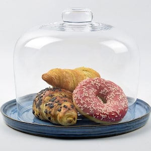 May include: A glass cake dome with a handle, covering a blue ceramic plate holding a croissant, a chocolate croissant, and a pink iced doughnut with sprinkles. The plate has a dark blue rim.