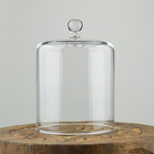 Może przedstawiać: Przezroczysta szklana klosz z okrągłym uchwytem na górze. Klosz stoi na drewnianej podstawie.