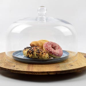 May include: A clear glass cloche with a round knob sits atop a wooden serving board. Inside, a blue plate holds a croissant, a chocolate croissant, and a pink frosted donut. The board has a natural wood grain.