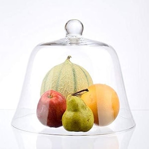 Könnte beinhalten: Eine transparente Glasglocke mit runder Basis und einem kuppelförmigen Deckel. In der Glocke befinden sich vier Früchte: eine Cantaloupe, eine Birne, ein Apfel und eine Zitrone.