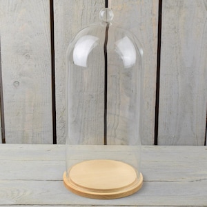 Może przedstawiać: Przezroczysty szklany klosz z zaokrąglonym wierzchołkiem i małym guzikiem. Stoi na okrągłej, jasnej drewnianej podstawie z ciemniejszą drewnianą obwódką. Tło stanowi jasnoszara drewniana powierzchnia.