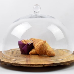 Op de afbeelding: Een glazen stolp met een ronde knop staat op een houten serveerplank. Binnenin worden een muffin in een paarse wikkel en twee croissants getoond. De houten plank heeft een natuurlijke, rustieke uitstraling.