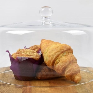 Pode incluir: Uma cúpula de vidro transparente para bolos com base redonda e pega pequena. Dentro, um muffin numa forma de papel roxa e um croissant dourado. A cúpula assenta numa superfície de madeira.