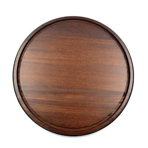 Può includere: Un vassoio rotondo in legno marrone scuro con un bordo rialzato e un'incisione vicino al bordo. La venatura del legno è visibile e il vassoio sembra essere fatto di un unico pezzo di legno. Il vassoio è progettato per servire cibo o esporre oggetti.