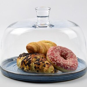 Puede incluir: Una cúpula de cristal transparente para pasteles con asa, que cubre un plato de cerámica azul con un cruasán de chocolate, un cruasán normal y un donut glaseado rosa con chispas. El plato tiene un borde azul oscuro.
