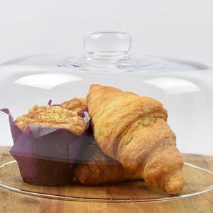 May include: A clear glass cake dome with a handle, displaying a muffin in a purple liner and two croissants on a wooden surface. The baked goods are golden brown, and the dome is transparent.