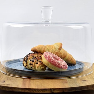 Puede incluir: Una cúpula de cristal transparente para pasteles con asa, que muestra productos horneados. Un plato azul contiene un pastel cubierto de chocolate, dos cruasanes y una rosquilla rosa. La cúpula se encuentra sobre una tabla de madera.