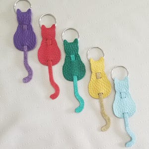 Könnte beinhalten: Fünf Leder-Schlüsselanhänger in verschiedenen Farben: lila, rot, grün, gelb und blau. Jeder Schlüsselanhänger zeigt eine Katzensilhouette mit einem langen Schwanz.