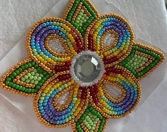 May include: A colorful beaded flower with a clear rhinestone center. The flower is made with small beads in shades of green, yellow, orange, red, purple, and blue.