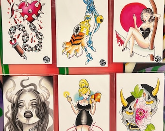 May include: A collection of six tattoo-style art prints. The prints feature various designs, including a heart with spikes, a tiger paw charm, a pin-up girl, a demonic woman, a woman with a book, and a Japanese demon mask. Each print is on a white background.