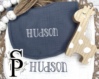May include: Two white muslin burp cloths with the name "Hudson" embroidered in white thread. The top cloth is a dark blue color. A wooden giraffe toy with white spots is on top of the cloths.