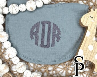 May include: A blue-green baby bib with a gray monogram. The bib is surrounded by white wooden beads and a wooden giraffe cutout. The bib has a rounded shape and is made of a soft, textured fabric.