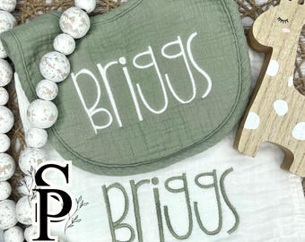 May include: Two white muslin burp cloths with the name "Briggs" embroidered in green and white thread. The cloths are displayed on a wooden surface with a wooden giraffe toy.