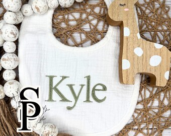 May include: White baby bib with the name "Kyle" embroidered in green. A wooden giraffe toy with white spots is next to the bib. The bib is surrounded by decorative wooden beads and a woven mat.
