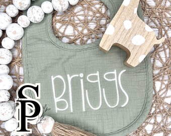 May include: A green muslin baby bib with the name "Briggs" embroidered in white thread. The bib is lying on a woven surface with a wooden giraffe toy.