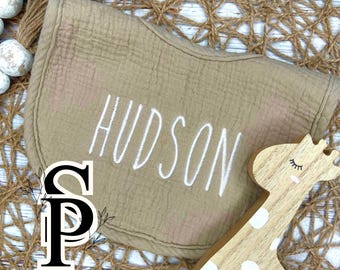 May include: A beige muslin burp cloth with the name "HUDSON" embroidered in white. The burp cloth is lying on a woven brown background with a wooden giraffe toy.