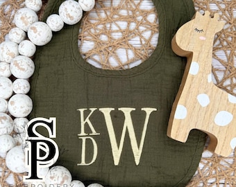 May include: An olive green baby bib with the embroidered initials "KDW" in a cream color. A wooden giraffe toy with white spots and a strand of white and brown wooden beads are next to the bib. The text "EMBROIDERY SHOP" is at the bottom.