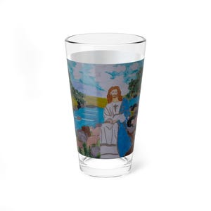 May include: A clear pint glass with a wraparound watercolour painting of a religious scene. The artwork shows a figure with long hair and a white robe, seated near water, with other figures and sheep. The background features a blue sky and green landscape.