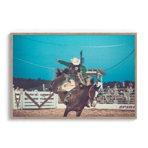 Op de afbeelding: Een cowboy in een groen shirt en een witte hoed rijdt een bruin paard in een rodeo-arena. Het paard bokt en de cowboy houdt zich vast. De arena is omgeven door een wit hek en er zijn mensen die toekijken op de achtergrond.