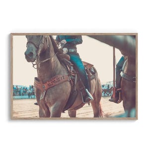 Könnte beinhalten: Ein braunes Pferd mit einem Reiter, der blaue Jeans und einen braunen Ledersattel trägt. Das Pferd befindet sich in einer Rodeo-Arena mit anderen Pferden und Reitern im Hintergrund.