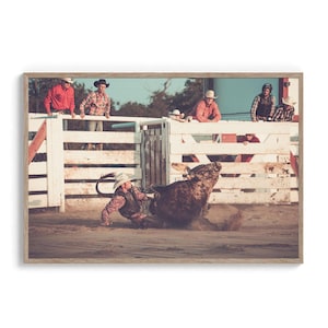 Op de afbeelding: Een rodeo cowboy wordt van een bruine stier gegooid in een arena met zand. De cowboy draagt een bruin vest, een witte hoed en een spijkerbroek. De stier bokst en schopt zand op. Toeschouwers kijken toe vanaf achter een wit houten hek.