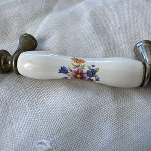 Puede incluir: Un tirador de armario vintage con un mango de cerámica blanca decorado con un estampado floral en tonos morado, azul y amarillo. Los herrajes metálicos tienen un aspecto envejecido y deslustrado.