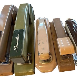 May include: A collection of vintage Swingline staplers in various colors, including brown, olive green, and tan. The staplers have a classic design with metal and plastic components. Some show signs of wear.