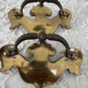 May include: Two antique brass drawer pulls with a dark bronze handle. The brass backplates have a decorative, ornate design. The pulls are set against a white lace background.