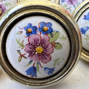 May include: Set of six vintage ceramic drawer pulls with a floral design. The pulls are white with pink, blue, and yellow flowers. They are set in a gold metal frame.