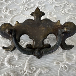 Op de afbeelding: Antiek messing ladetrekker met een decoratief, sierlijk ontwerp. De trekker heeft een lusvormige handgreep en is in een verweerde afwerking.