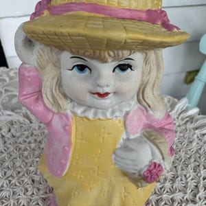 May include: A ceramic figurine of a girl wearing a yellow dress, pink jacket, and a straw hat with a pink ribbon. She is holding a basket with a pink flower.