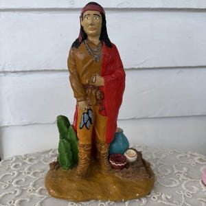 May include: A hand-painted figurine of a Native American man. He wears a brown shirt, orange pants, and a red shawl. The figurine stands on a brown base with a cactus, a blue vase, and small pottery. The figure has long black hair and a red headband.