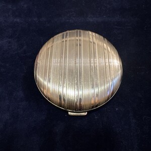 May include: A round, gold-colored compact with a ridged design. The compact has a hinged lid and a small latch.