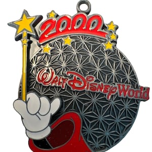 Peut inclure: Ornement commémoratif Walt Disney World 2000. L'ornement présente un globe stylisé avec le texte "Walt Disney World" en rouge. Une main gantée tient une baguette jaune avec une étoile. L'année "2000" est en rouge.