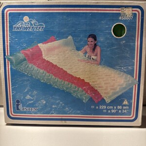 May include: A colorful inflatable pool float with a green, pink, and white striped design. The float is in a swimming pool and a person is laying on it. The box has the text "The Wet Set" and "INTEX" on it. The float is 90 inches by 34 inches.